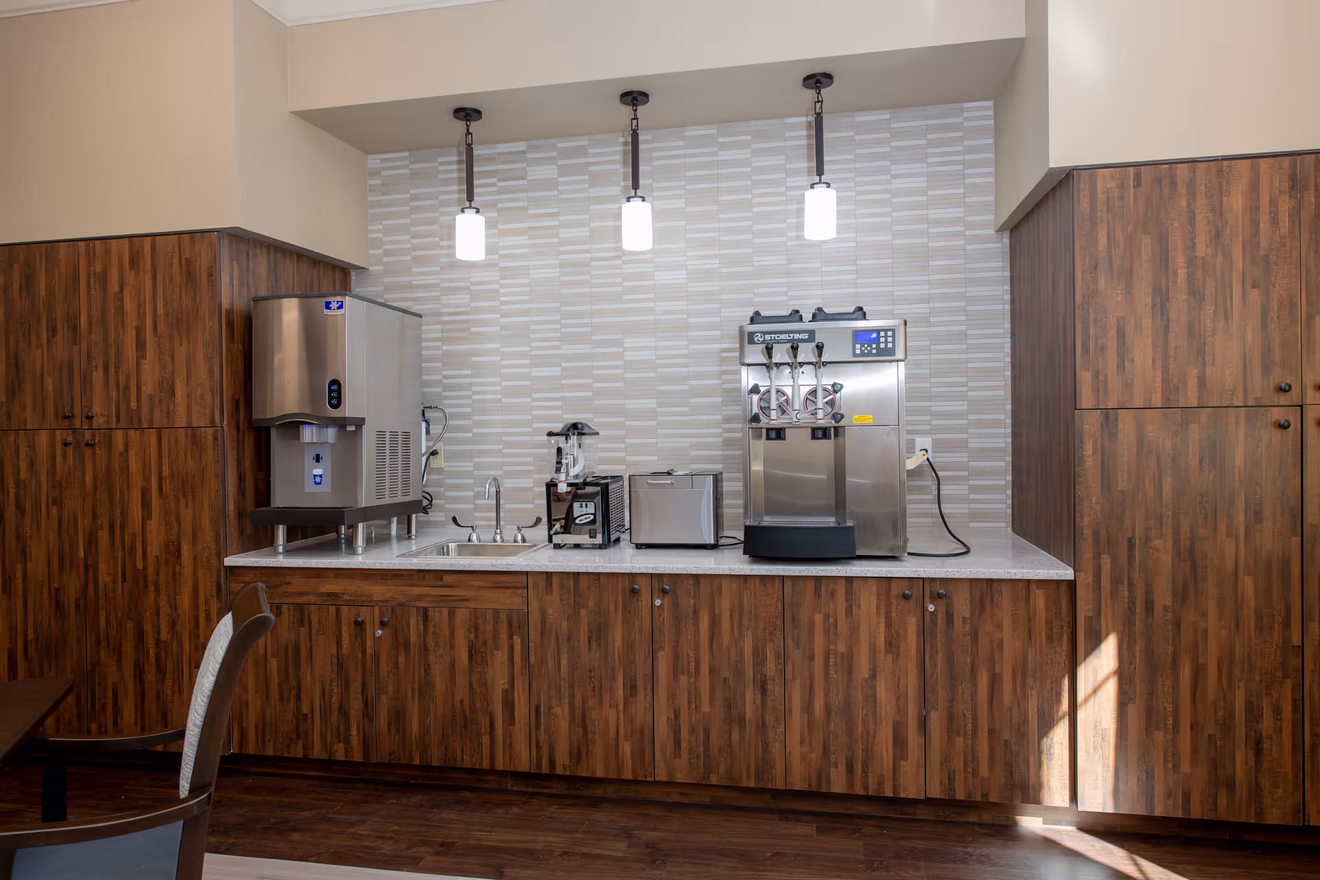 A kitchenette area with wooden cabinets, a countertop with a sink, a commercial ice machine, a blender, a toaster, and a soft serve ice cream machine. Three pendant lights hang above the countertop, and the backsplash features a light gray and white tile pattern.