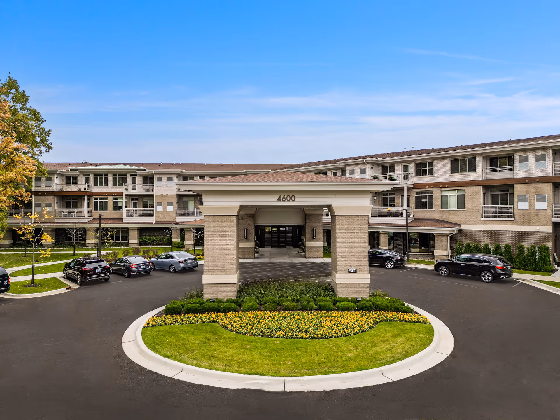Front exterior view of a modern three-story senior living facility with a covered entrance, circular driveway, parked cars, landscaped flower beds, and green lawns under a clear blue sky.