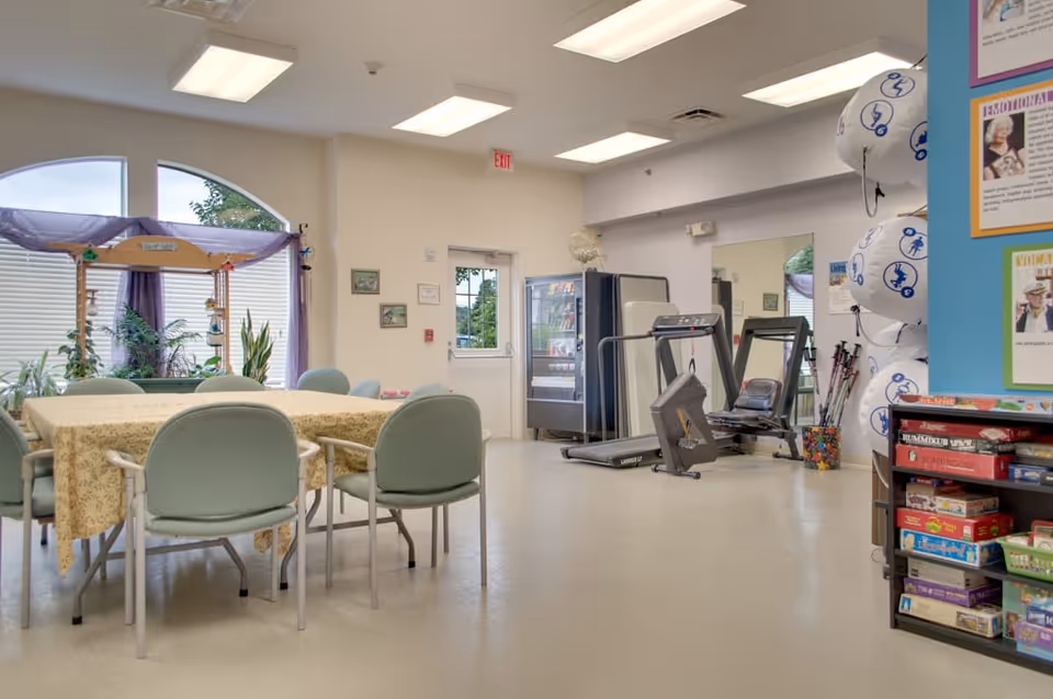 A bright and spacious common area in a senior living facility featuring a table with a floral tablecloth surrounded by green cushioned chairs. In the background, there is exercise equipment including a treadmill and a recumbent bike, a vending machine, and a large mirror on the wall. To the right, there is a shelf filled with board games and balloons with disability symbols. Large windows with purple curtains and several potted plants add natural light and greenery to the room.