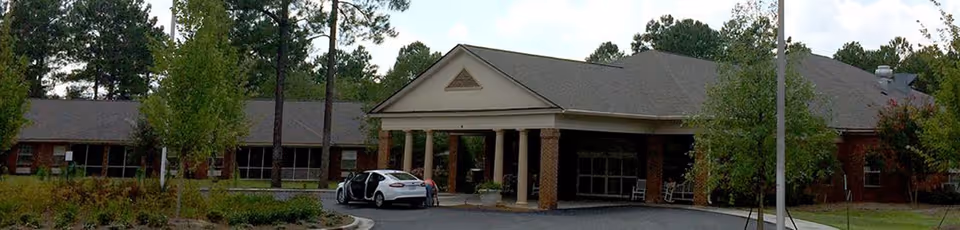 Front exterior view of Legacy Village at Park Regency, showing a single-story brick building with a covered entrance supported by columns, surrounded by trees and landscaping, with a white car parked near the entrance.