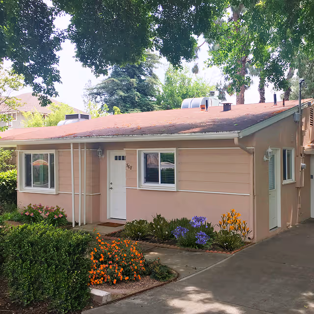Single-story beige building with a red roof, white doors and windows, surrounded by green bushes and colorful flowers, with large trees providing shade.