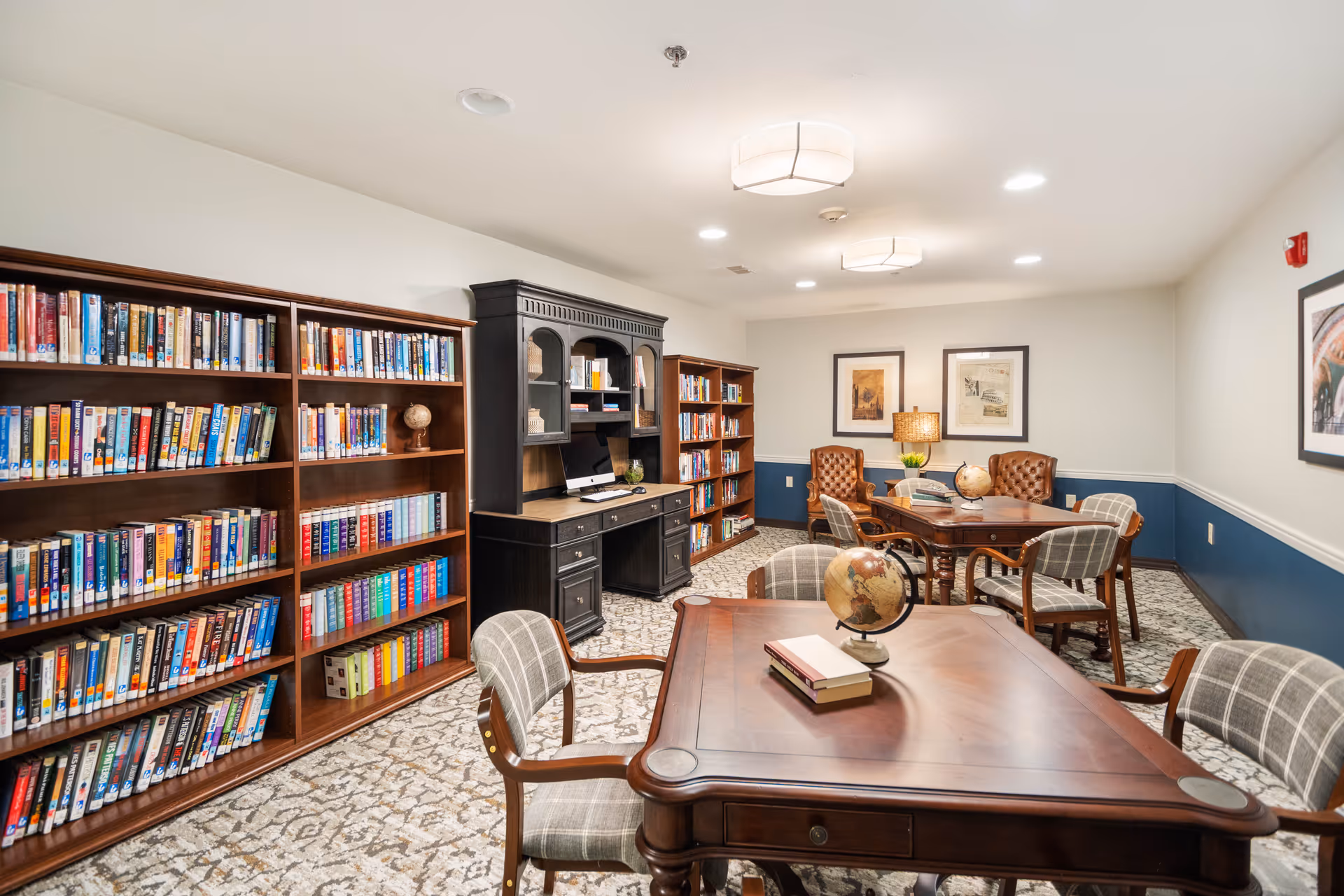A well-lit library or reading room with wooden bookshelves filled with books, a black desk with a computer, and several wooden tables with chairs. The tables have globes and books on them. The walls are painted white with a blue lower half, and framed artwork is hung on the walls.