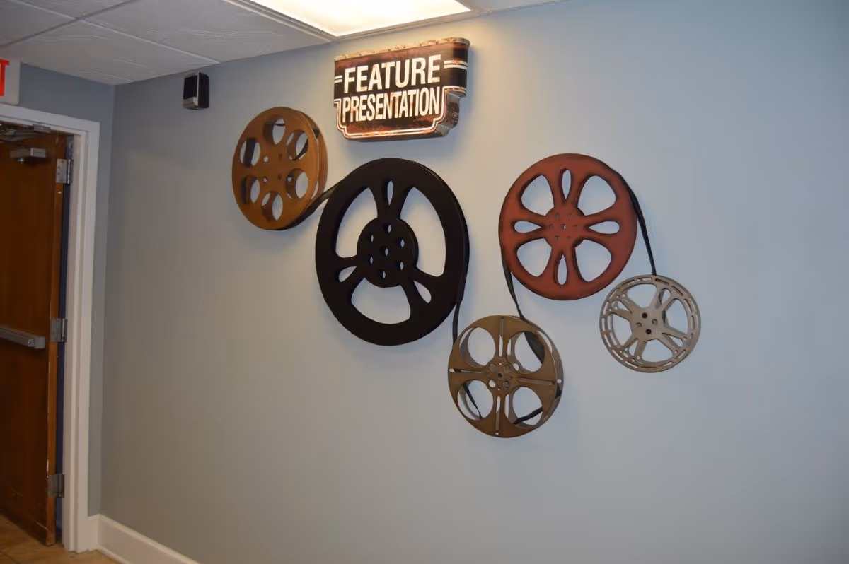 Wall decoration featuring five vintage film reels of different sizes connected by a black strip, with a sign above them that reads 'FEATURE PRESENTATION' in white letters on a brown background. The wall is painted light blue, and there is a wooden door with metal hinges on the left side.