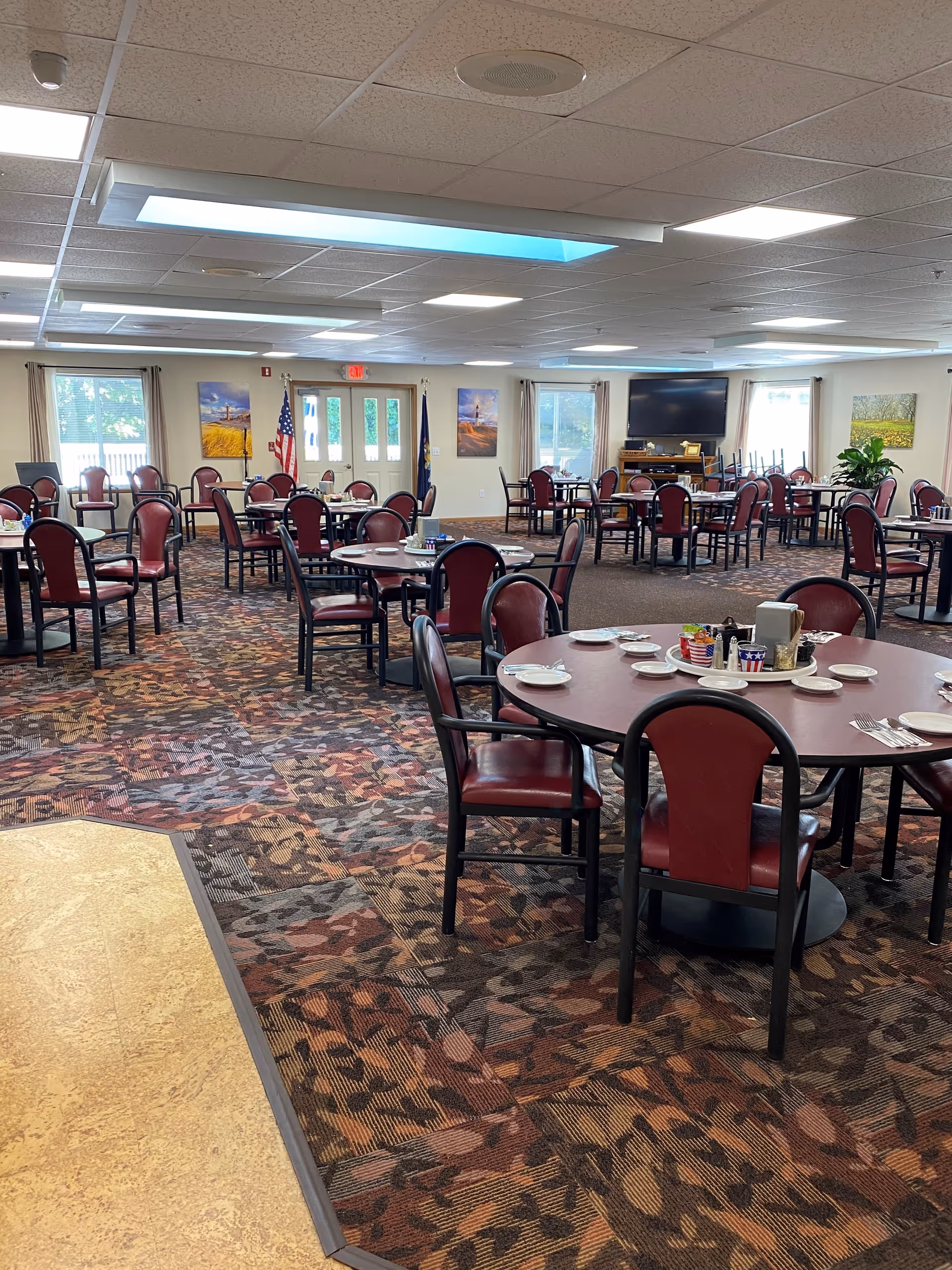A spacious dining room with multiple round tables and red cushioned chairs arranged neatly. Each table is set with plates, utensils, and condiments. The room has patterned carpet flooring, large windows with curtains, and a television mounted on the far wall. There are also two flags near the exit door and framed pictures on the walls.