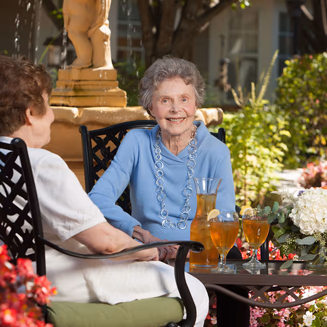 Two elderly women sitting outdoors at a table with iced tea drinks, surrounded by flowers and greenery, with a stone fountain in the background. One woman is smiling and wearing a light blue sweater and a large necklace.