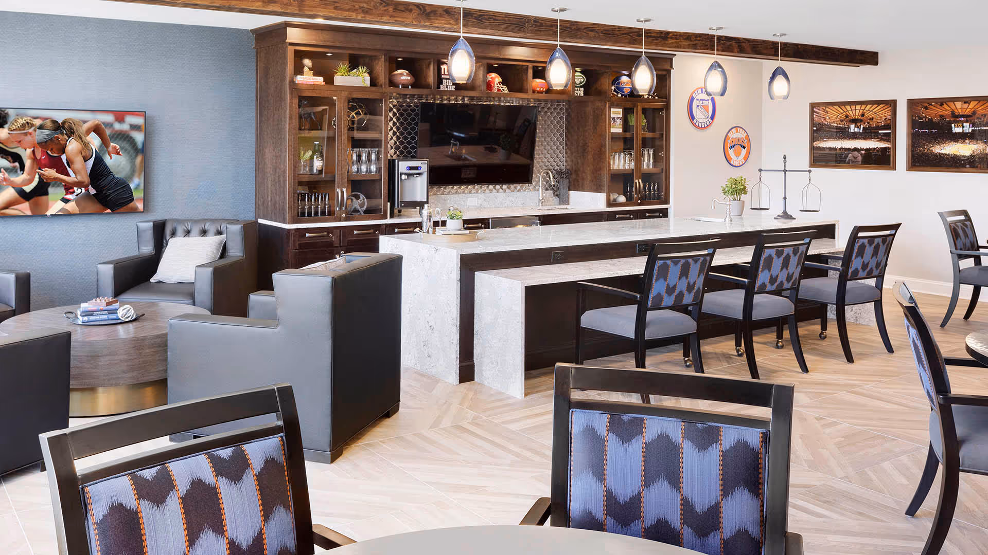 A modern common area with a kitchen and lounge seating. The kitchen features a marble countertop island with bar stools, dark wood cabinetry, a built-in TV, and pendant lighting. The lounge area has gray armchairs and a round coffee table. The walls are decorated with sports-themed pictures and memorabilia.