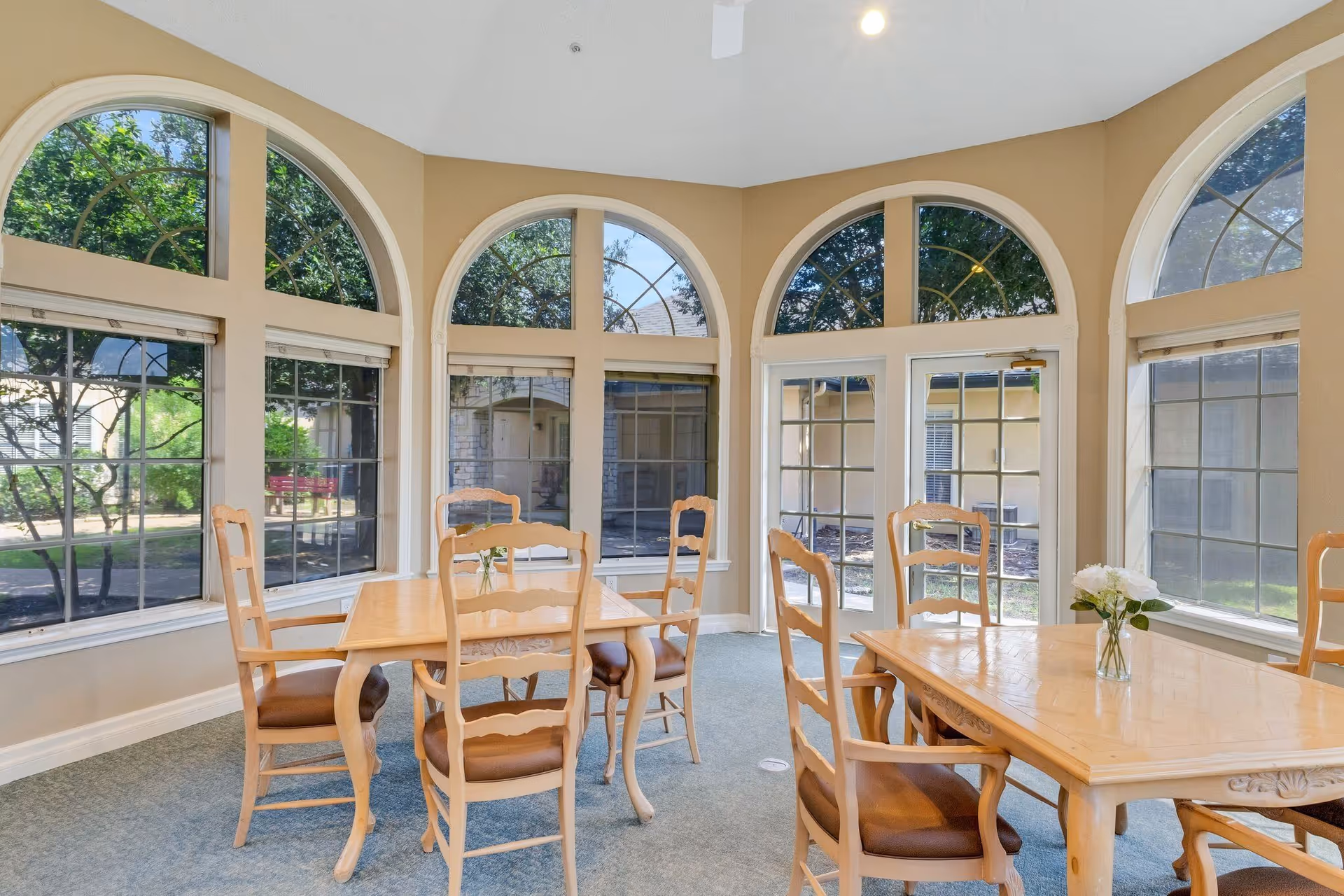 A bright dining room with large arched windows allowing natural light to fill the space. The room contains two wooden dining tables, each surrounded by wooden chairs with cushioned seats. A small vase with white flowers is placed on one of the tables. Outside the windows, greenery and parts of the building exterior are visible.