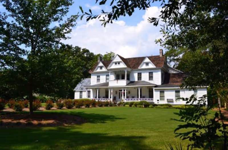 A large white two-story house with a brown shingled roof, surrounded by green grass, trees, and shrubs under a partly cloudy sky.