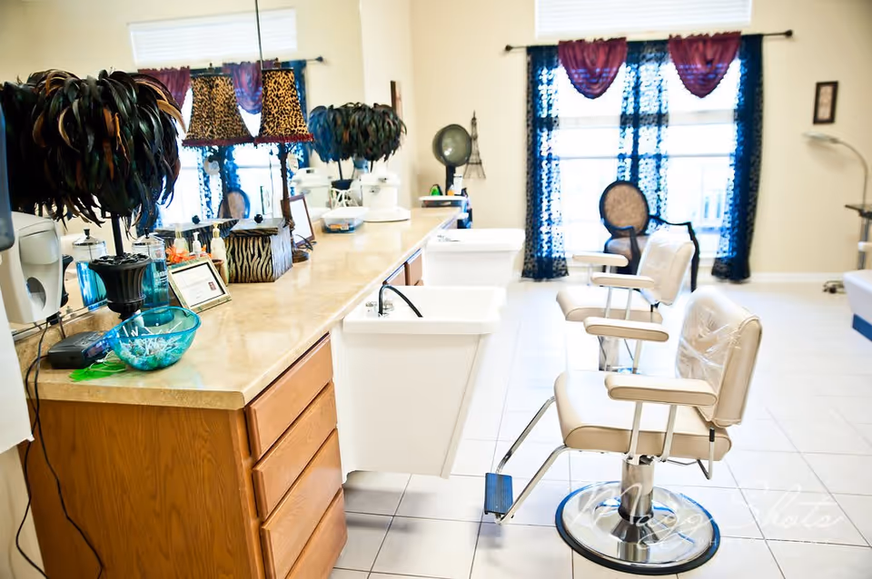 Interior view of a salon area with two white salon chairs in front of sinks, a long countertop with various decorative items including feathered lamps and a bowl with cotton swabs, and a large mirror on the wall. The room has tiled floors, a window with sheer black curtains and purple valances, and additional seating in the background.