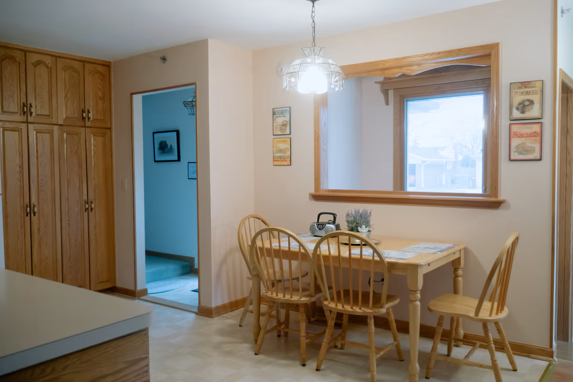 Dining nook with a wooden table and four matching chairs beneath a hanging light, oak cabinets to the left and a pass-through window to the right.