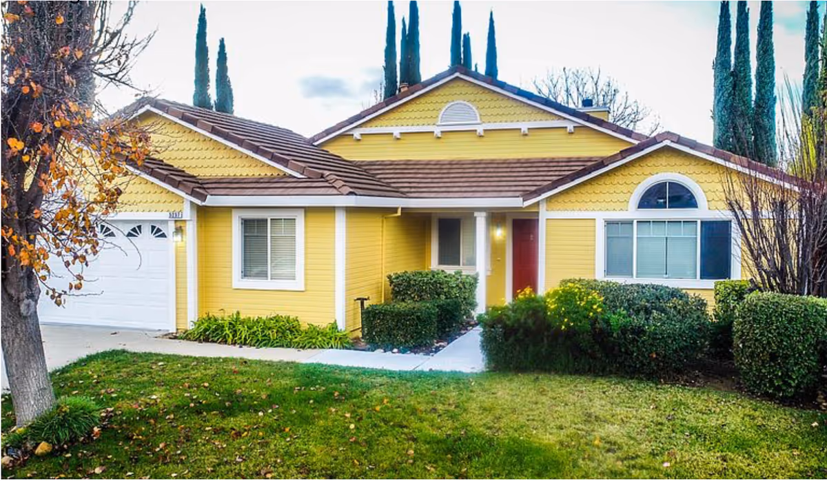 Front exterior of a yellow single-story house with a garage, red front door, and landscaped lawn and shrubs.