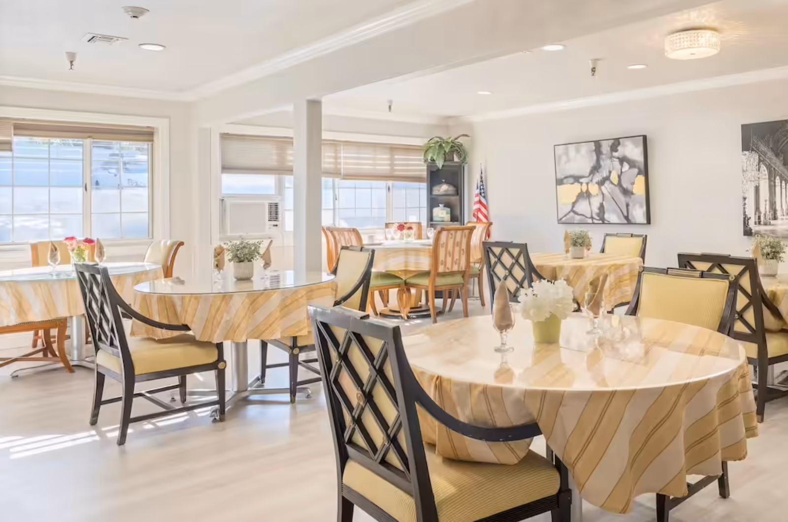Bright dining room with round tables draped in yellow-striped tablecloths, chairs, floral centerpieces, and wall art.