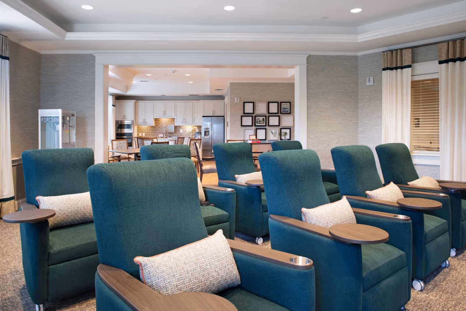 Teal reclining armchairs with attached side tables arranged in a communal lounge, with a popcorn machine and an open kitchen/dining area visible in the background.