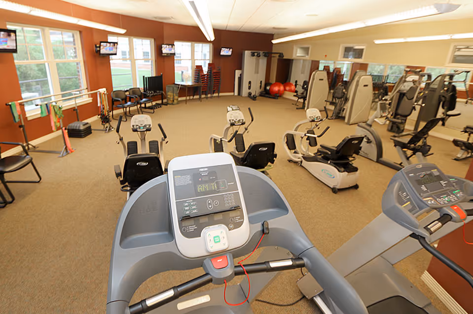 Fitness room with treadmills in the foreground, recumbent bikes and other exercise equipment against mirrored walls and windows.