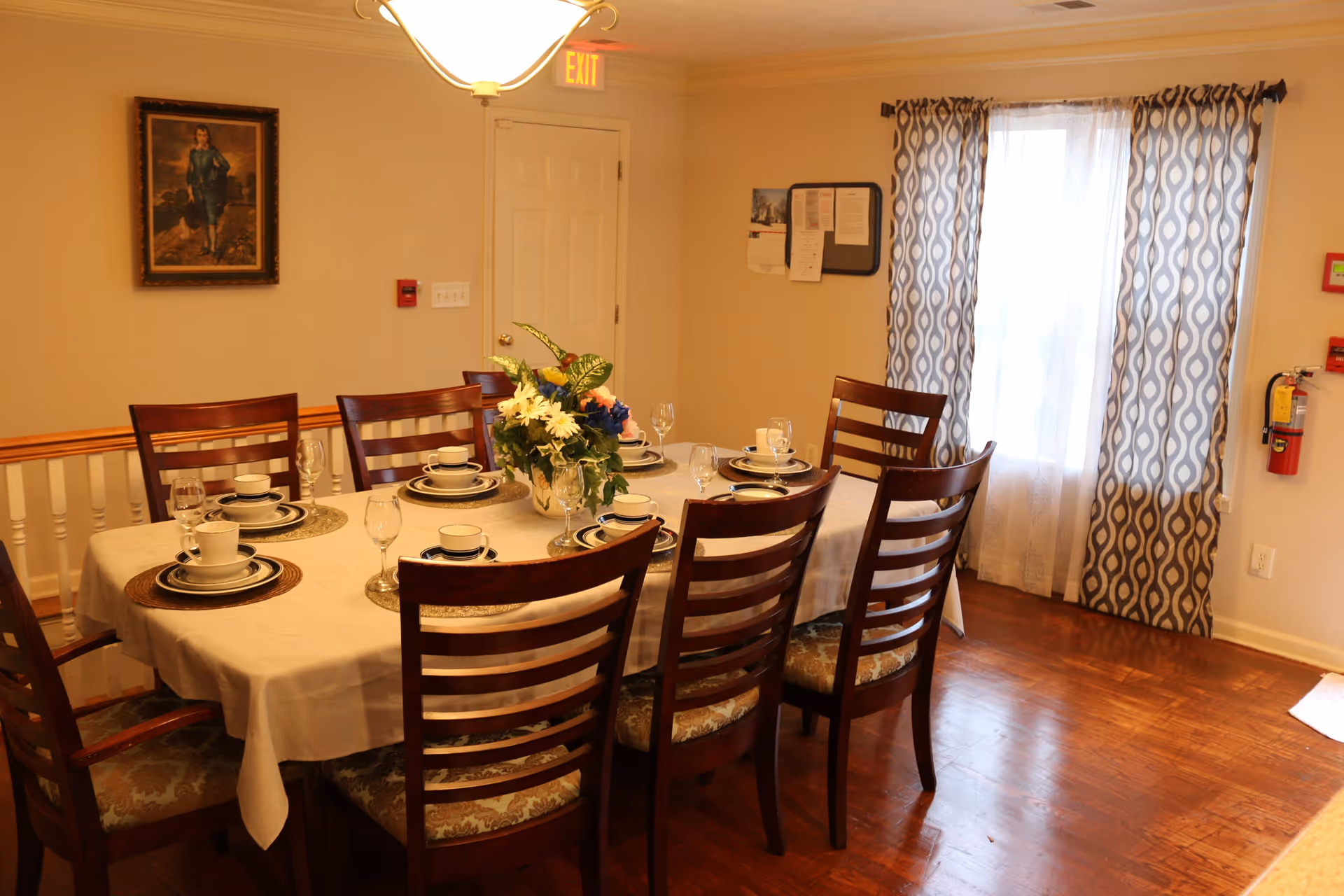 A dining room with a rectangular table covered with a white tablecloth set for eight people. The table has matching dark wooden chairs with patterned cushions. Each place setting includes a plate, bowl, cup, and glass. A floral centerpiece decorates the middle of the table. The room has wooden flooring, a window with patterned curtains, a painting on the wall, and an exit sign above a door.