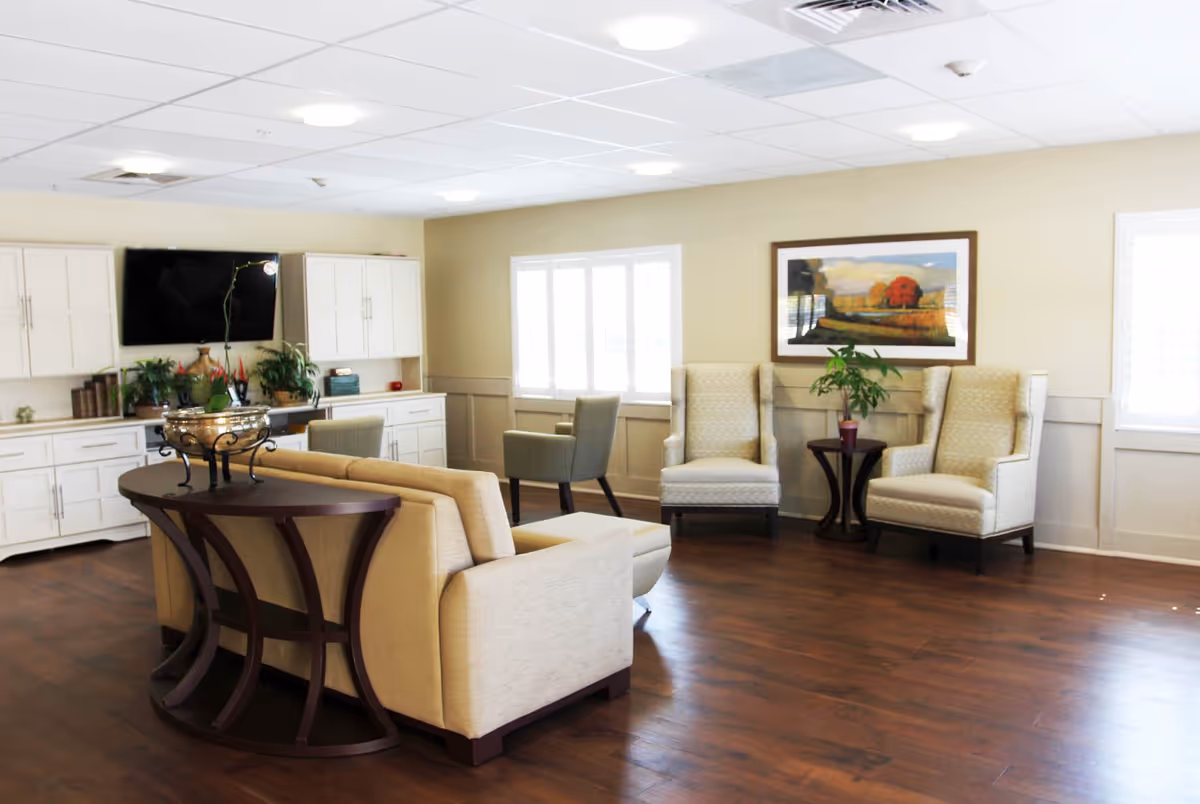 A bright and spacious living room area in The Lodge Health & Rehabilitation Center featuring a beige sofa with a dark wooden console table behind it, two beige armchairs with a small round table and a plant between them, a wall-mounted flat-screen TV, white cabinets, and a framed landscape painting on the wall. The room has wooden flooring and large windows letting in natural light.