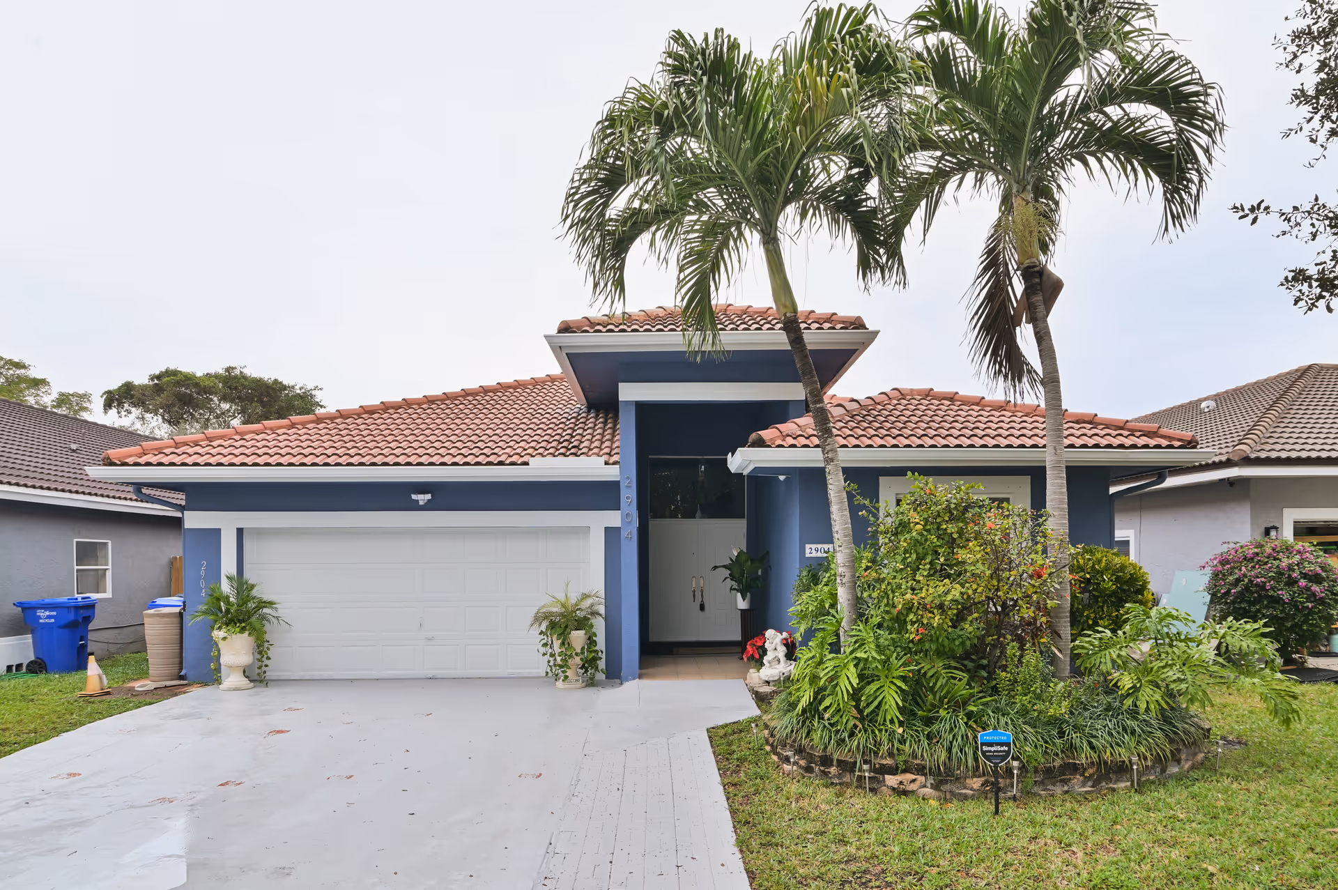 Front exterior view of a single-story house with a tiled roof, blue walls, a white garage door, and a driveway. There are two palm trees and various shrubs and plants in a landscaped garden area near the entrance.
