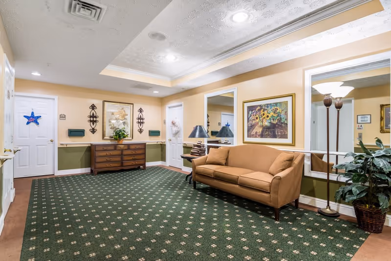 A cozy interior common area with a green patterned carpet, a brown sofa with two cushions, a wooden side table with a lamp, a tall floor lamp, a large potted plant, and framed artwork on the beige walls. There are two white doors, a wooden dresser with decorative items, and two large mirrors on the wall reflecting the room.