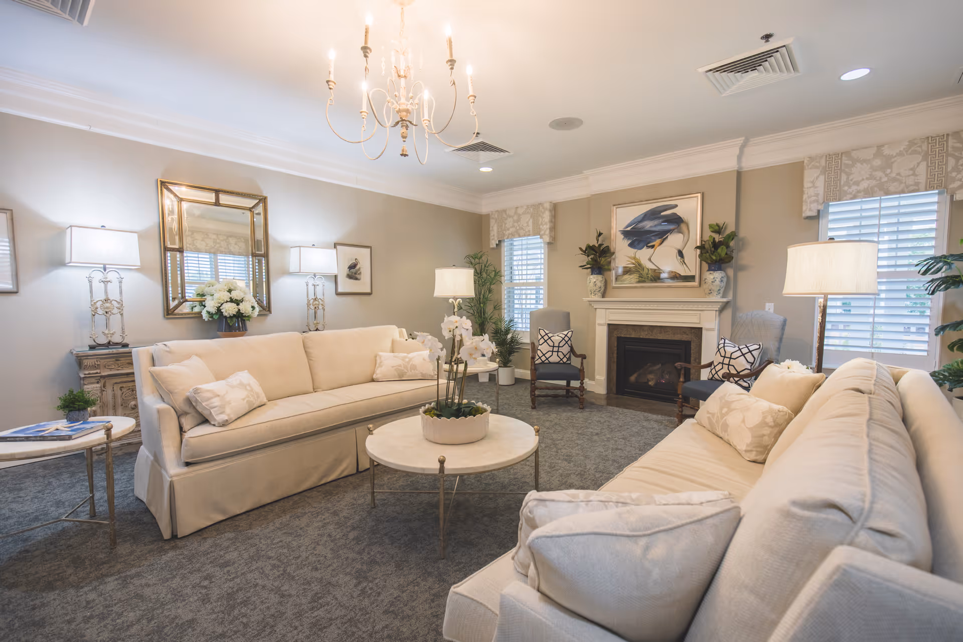 A cozy living room with two beige sofas facing each other, a round coffee table with a plant in the center, two armchairs near a fireplace, decorative lamps, a chandelier, framed artwork, and windows with white blinds and patterned valances.