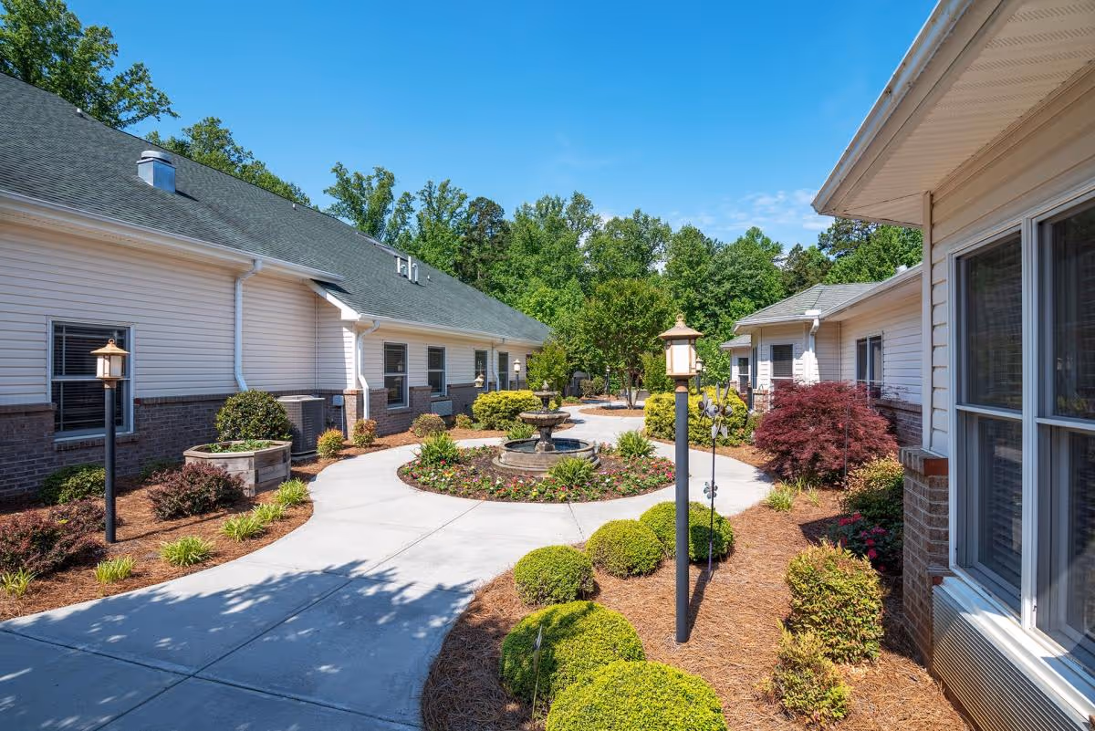 Outdoor courtyard area at Brookstone of Clemmons featuring a circular walkway around a central fountain, surrounded by well-maintained bushes, plants, and trees with buildings on either side under a clear blue sky.