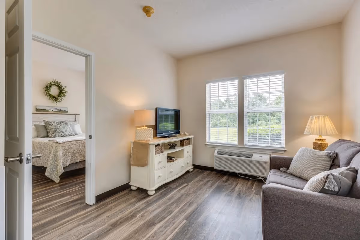 A cozy living room with a gray sofa adorned with pillows, a white TV stand holding a flat-screen television and a lamp, and two large windows with white blinds letting in natural light. To the left, an open door reveals a bedroom with a bed decorated with patterned bedding and a green wreath hanging above the headboard.