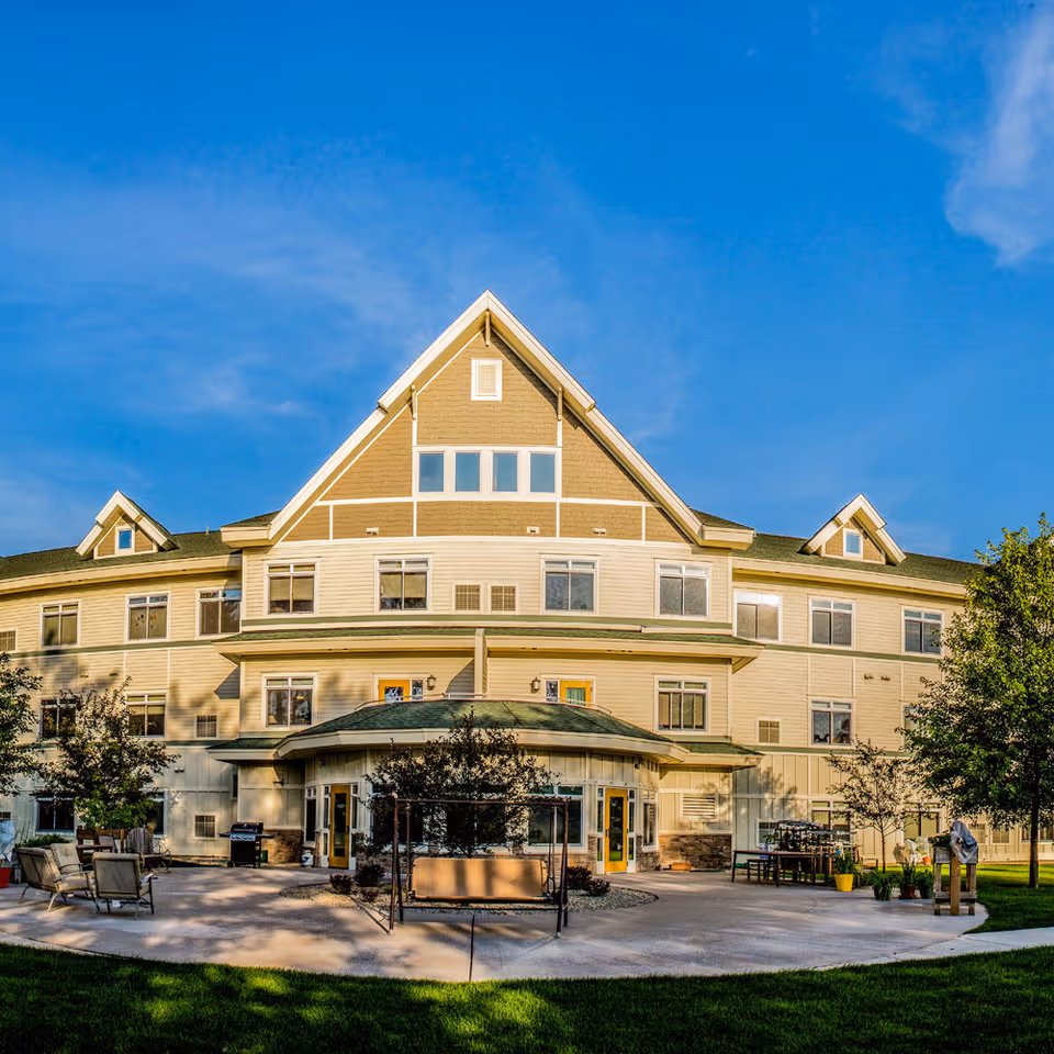 Front exterior of a multi-story beige assisted living building with a patio and outdoor seating under a clear blue sky.