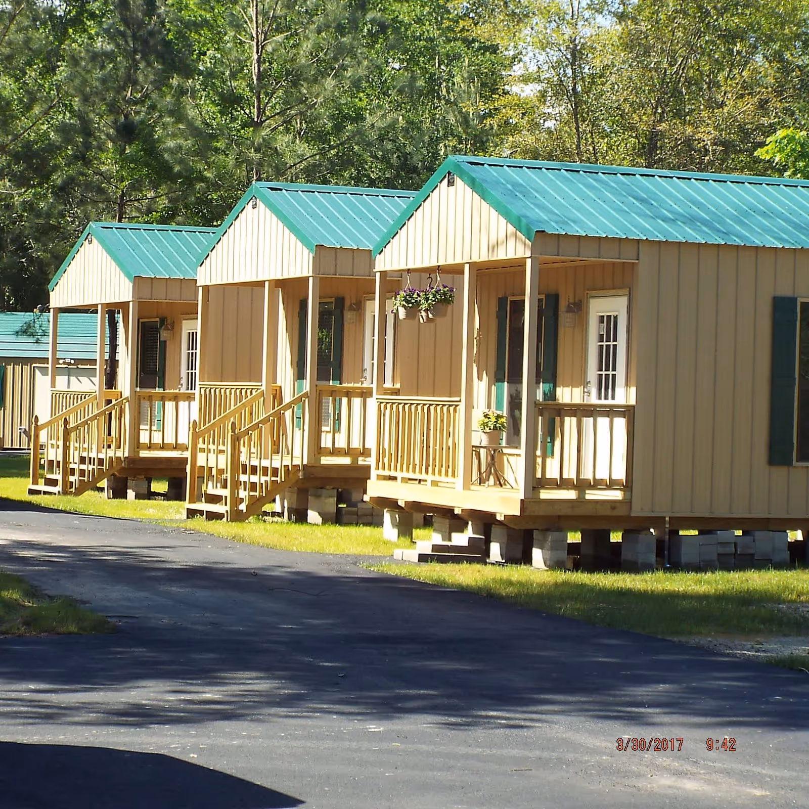 Row of small tan cabins with green metal roofs and front porches along a paved road surrounded by trees.