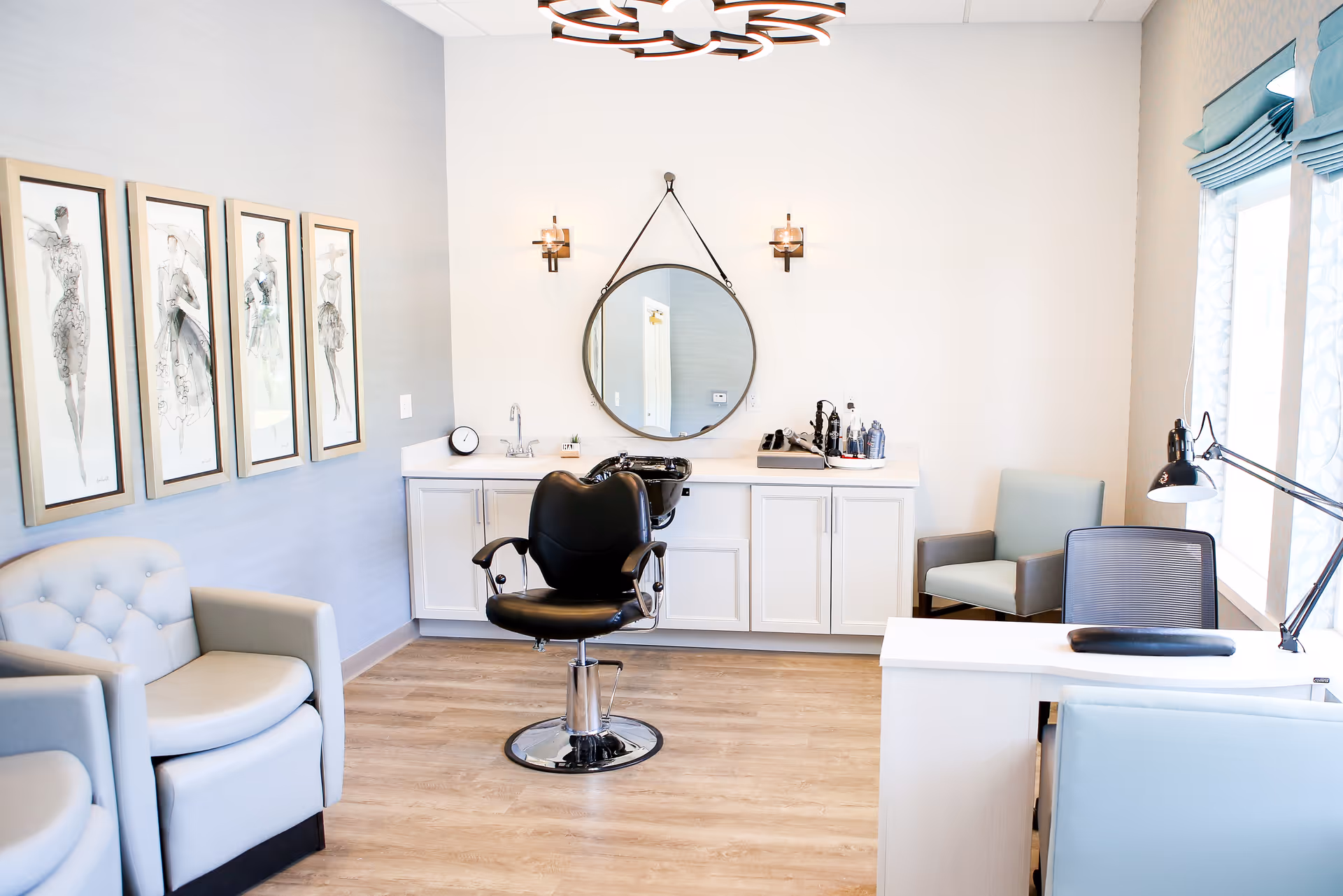 Bright salon-style interior with a black styling chair in front of a round mirror, white cabinets, and cushioned seating.