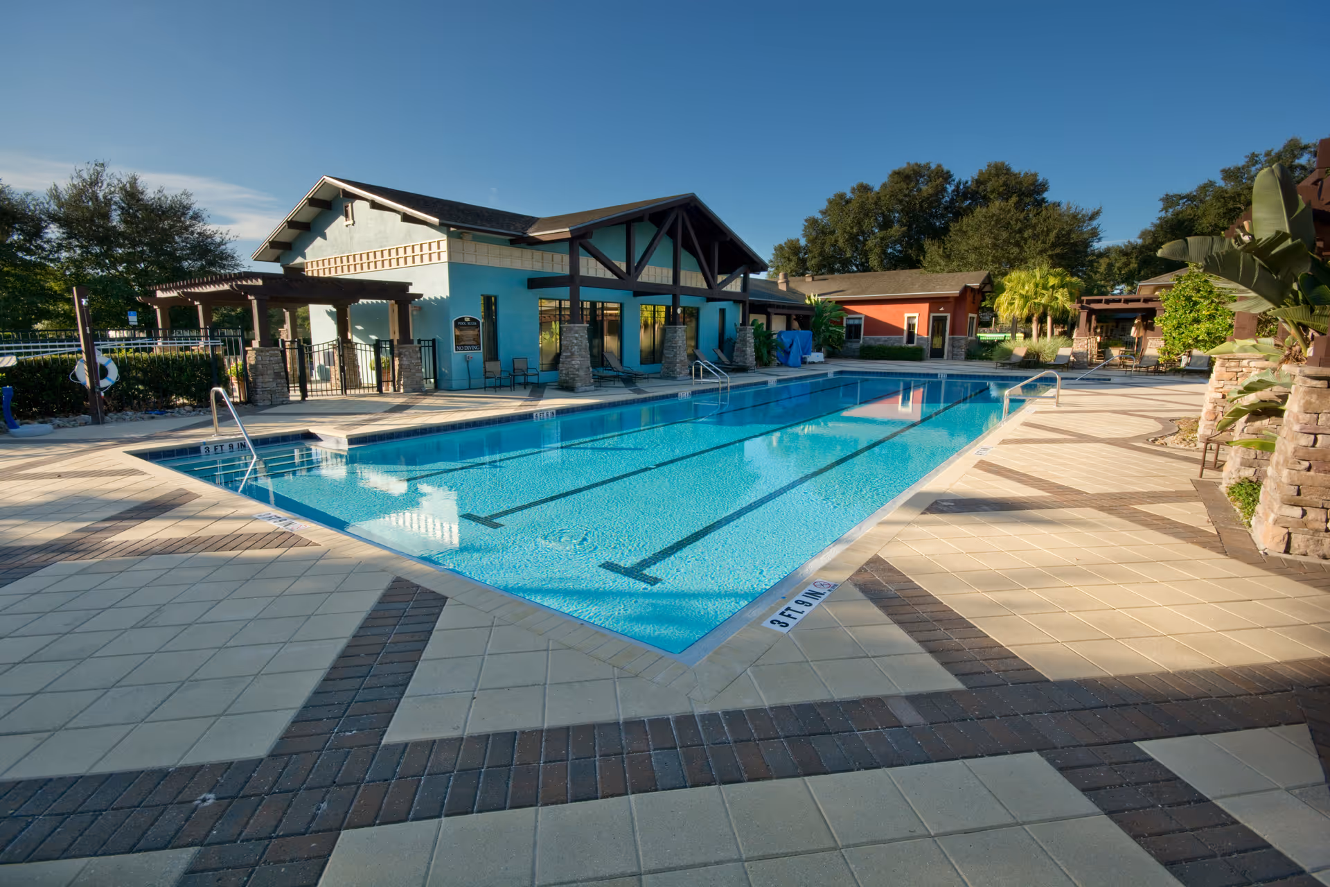 Outdoor swimming pool with clear blue water surrounded by a tiled deck area. There are buildings and shaded seating areas around the pool, with trees and greenery in the background under a clear blue sky.
