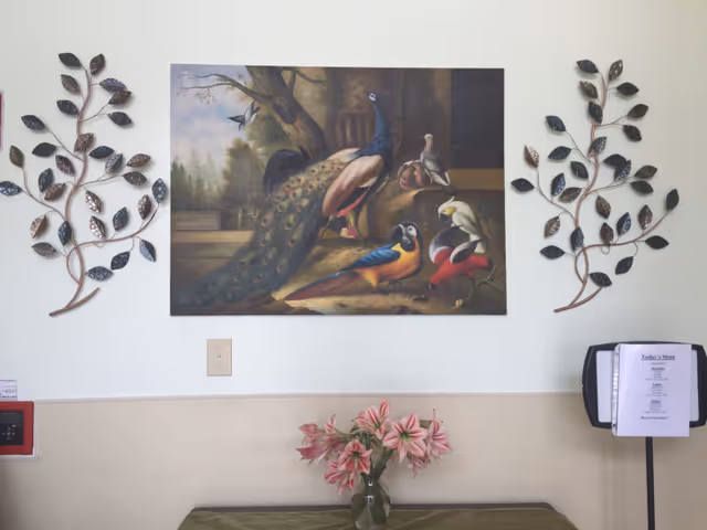 A wall with a large painting of birds flanked by metal leaf decorations above a table holding a vase of pink flowers and a menu stand.