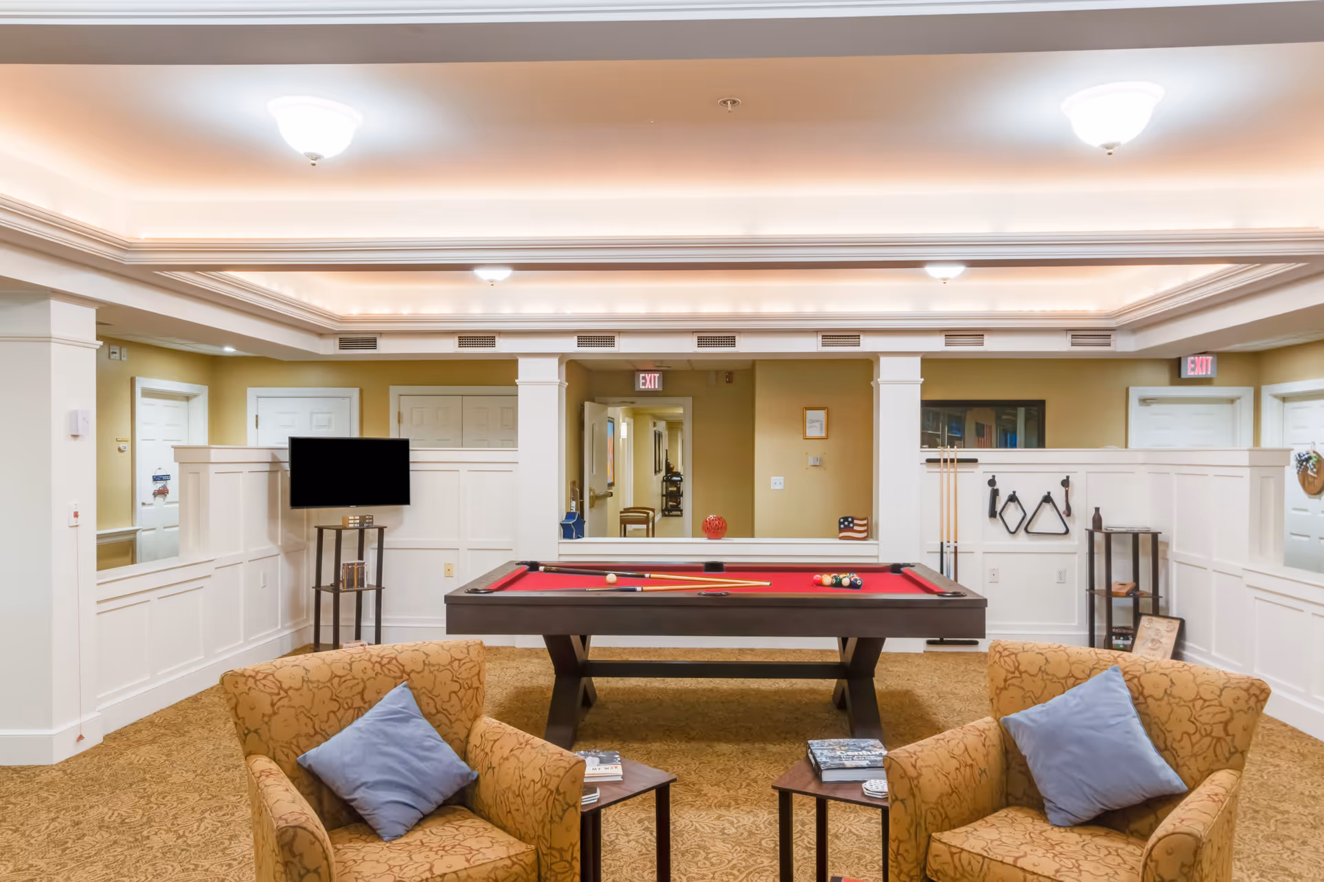 A cozy recreational room featuring a red felt pool table in the center with pool cues and balls arranged on it. Two patterned armchairs with blue pillows face the pool table, each accompanied by a small wooden side table holding books and a coaster. The room has beige walls with white wainscoting, recessed lighting in the ceiling, and a small TV mounted on a stand to the left. There are doors and hallway visible in the background.