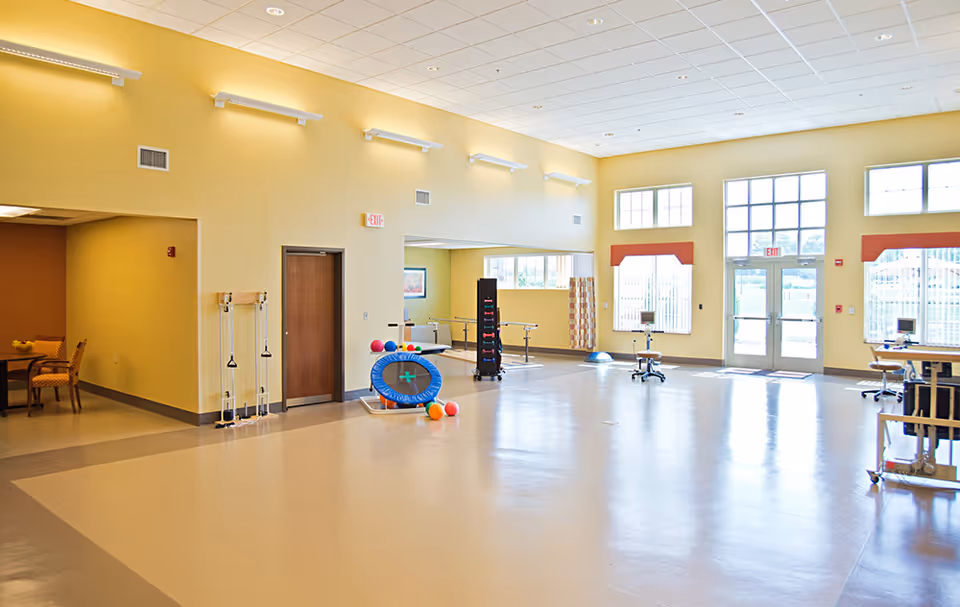 A spacious, well-lit room with large windows and double glass doors letting in natural light. The room has light yellow walls and a beige floor with a darker border. There is exercise equipment including a small trampoline, colorful balls, resistance bands, and parallel bars. To the left, there is a small seating area with a table and chairs. The ceiling has recessed lighting and a grid pattern. The room appears to be designed for physical therapy or exercise activities.