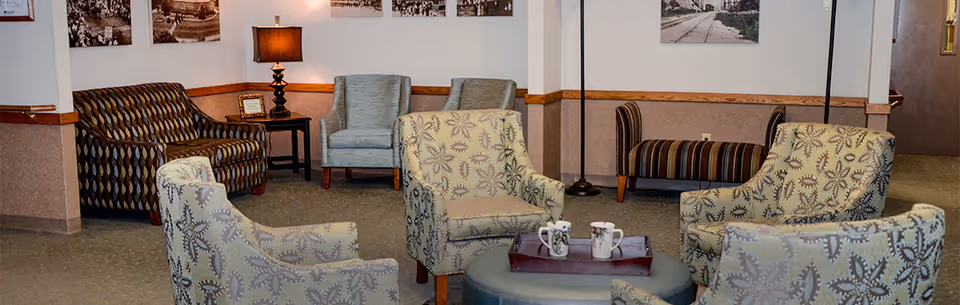 A cozy seating area in a senior living facility with multiple upholstered armchairs arranged around a round ottoman with a tray holding two mugs. The walls are decorated with framed black and white photographs, and there is a table lamp on a side table next to a patterned armchair. The space has a warm and inviting atmosphere.