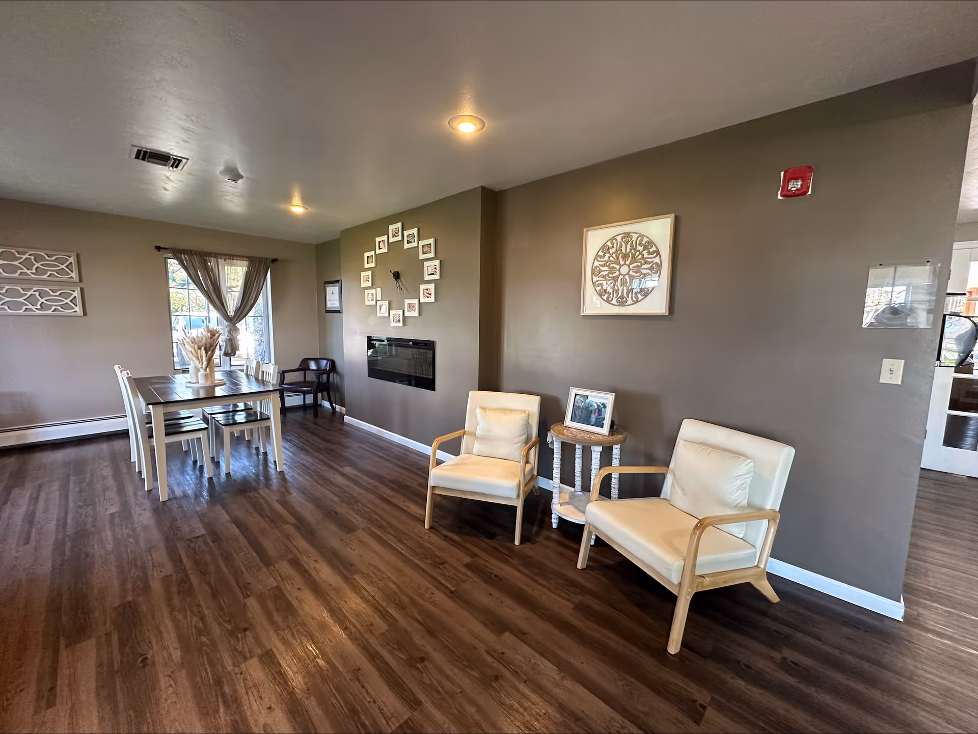 A cozy interior room with wooden flooring featuring a dining table with six chairs near a window with sheer curtains. On the right side, there are two cream-colored armchairs with wooden frames separated by a small round table holding a framed photo. The wall behind the chairs is decorated with a framed circular artwork and a clock made of small framed pictures arranged in a circle above a modern wall-mounted electric fireplace.