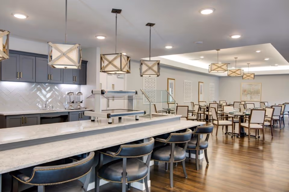 A spacious dining area with a long marble counter and black cushioned bar stools in the foreground. Behind the counter is a kitchen area with grey cabinets, a white herringbone tile backsplash, and two beverage dispensers. The dining area has multiple tables and chairs arranged neatly, with modern pendant lights hanging from the ceiling and wooden flooring throughout.
