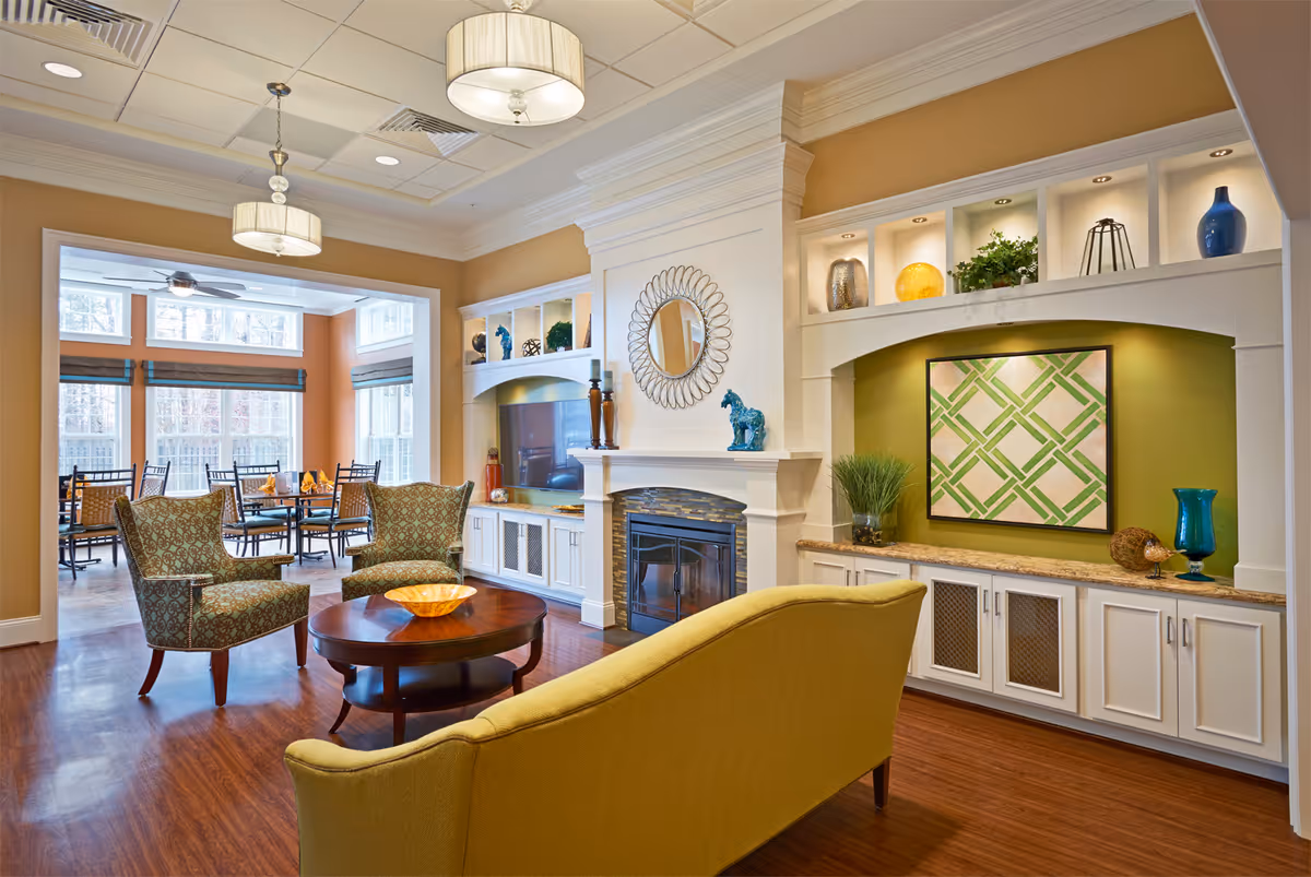 A cozy and well-lit living room area in Brandermill Woods Retirement facility featuring a yellow sofa, two patterned armchairs, a round wooden coffee table with a decorative bowl, a fireplace with a round mirror above it, built-in shelves with decorative items, and a dining area with tables and chairs visible through a large open doorway.