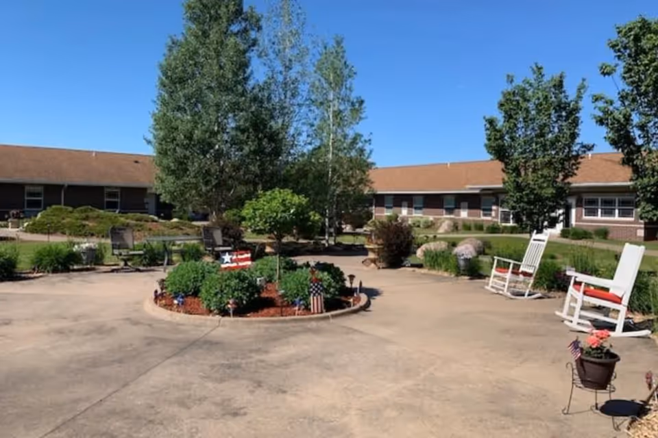 Sunny courtyard with a circular planted bed, rocking chairs, and a single-story building in the background under a clear blue sky.