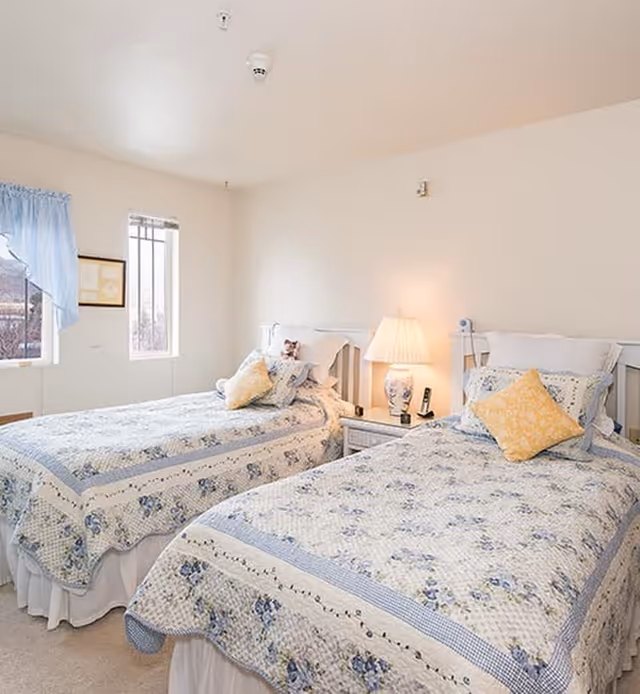 A bright bedroom with two twin beds, each covered with a floral quilt in shades of blue and white. Each bed has a yellow decorative pillow. Between the beds is a white nightstand with a lamp and a phone. The room has light-colored walls and two windows with blue curtains letting in natural light.