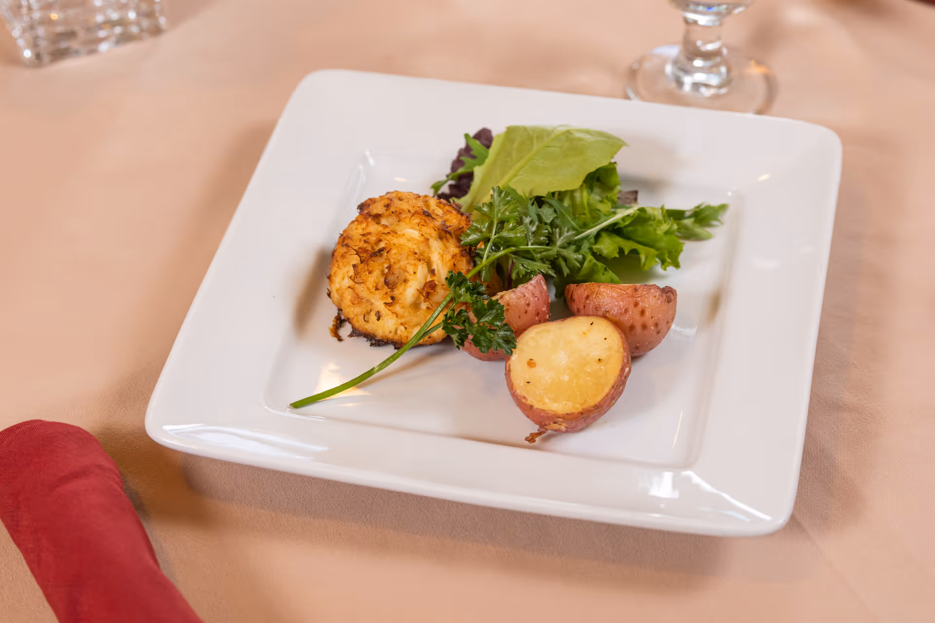 A white square plate with a small salad, roasted red potatoes, and a crab cake on a beige tablecloth.