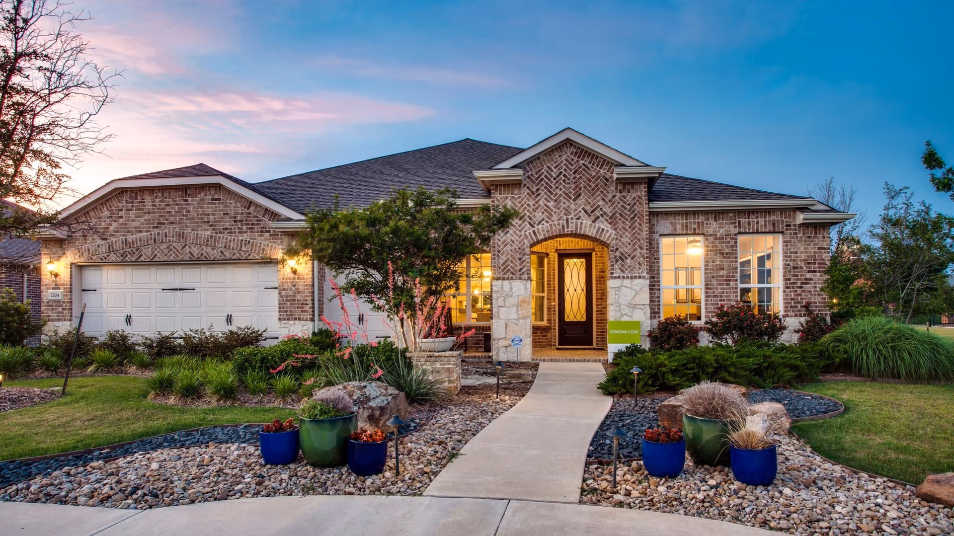 Front exterior view of a single-story brick house with a two-car garage, a pathway leading to a dark wooden front door with glass panels, and landscaped garden beds with rocks, plants, and potted flowers on either side. The house is illuminated with warm lights during dusk under a partly cloudy sky.