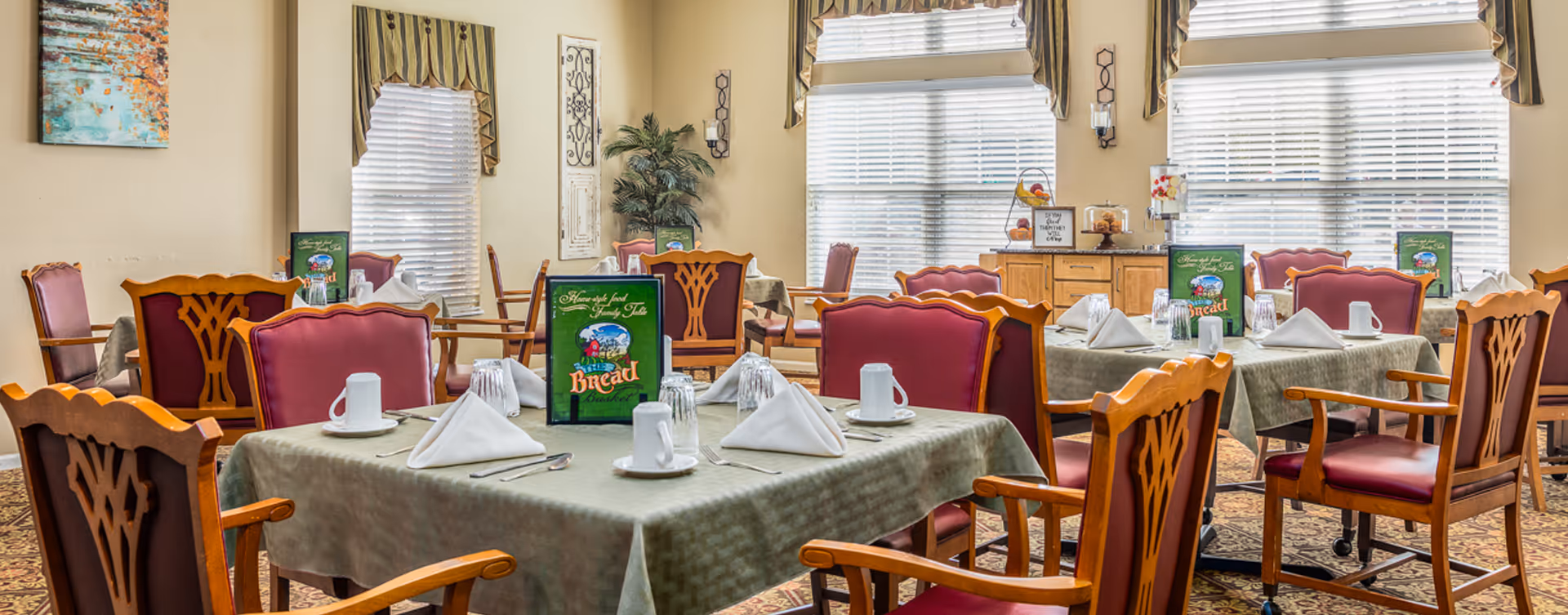 A dining room with multiple tables covered with green tablecloths, each set with white napkins, cups, glasses, and silverware. The chairs have wooden frames with red cushioned seats and backs. Large windows with blinds and valances allow natural light to fill the room. There is a sideboard with a fruit basket, pastries, and a beverage dispenser near the windows. The room has beige walls, decorative wall hangings, and a patterned carpet.
