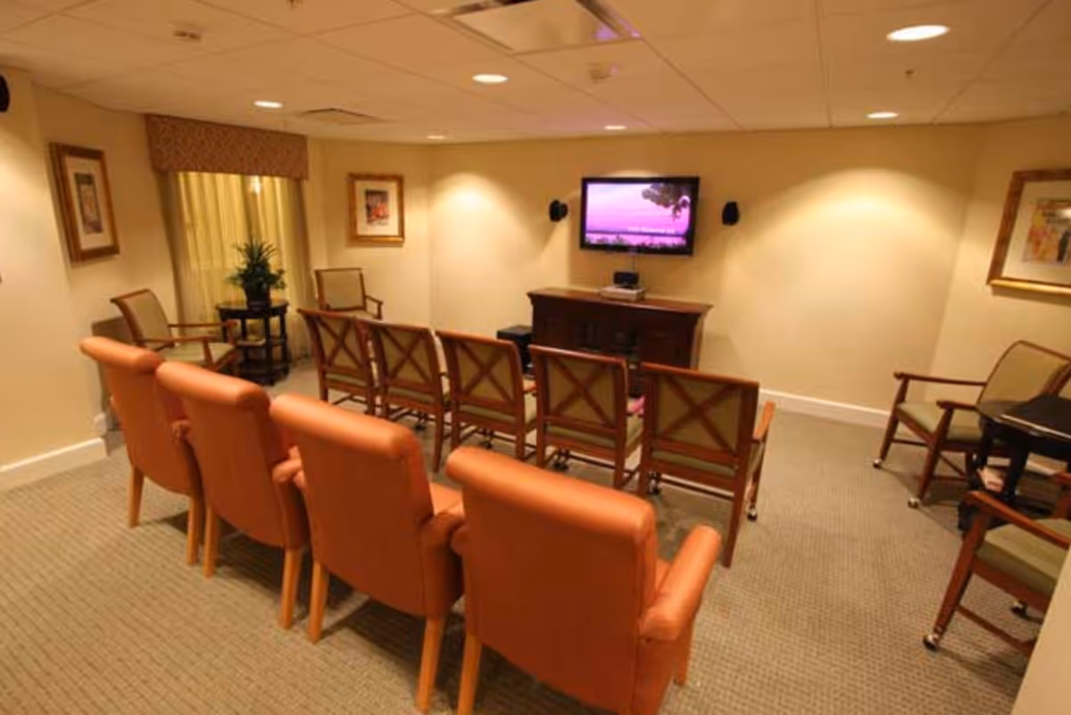 A small media room with two rows of chairs facing a wall-mounted flat screen TV. The front row has four cushioned armchairs in a warm brown color, and the back row has six wooden chairs with cushioned seats. The room has beige walls, carpeted floor, framed artwork on the walls, and a small table with a plant near a window with curtains.