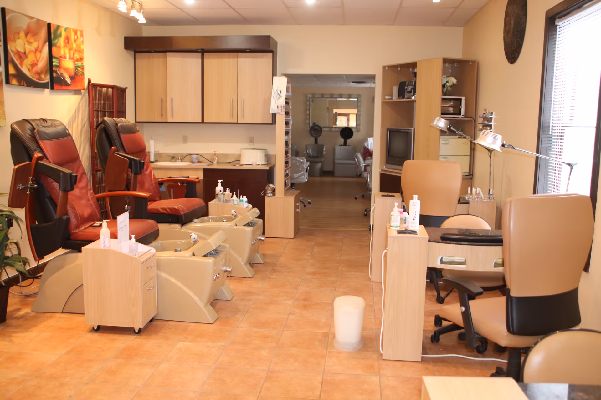 Interior view of a salon area in a senior living facility with two pedicure chairs on the left, manicure stations with chairs on the right, and a doorway leading to another room with hair drying chairs in the background. The room has tiled floors, beige walls, and wooden cabinets.