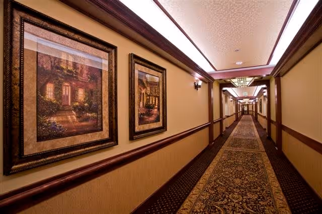 A long, well-lit hallway in a retirement community with patterned carpet and beige walls. The walls are decorated with framed paintings of houses and gardens. The ceiling has recessed lighting and decorative molding.