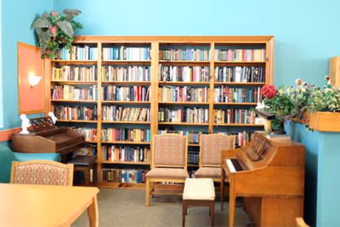 A cozy room with a large wooden bookshelf filled with books against a turquoise wall. In front of the bookshelf are two patterned chairs and a small cushioned stool. To the right, there is a wooden piano with a floral arrangement on top. On the left side, part of a wooden table and a chair are visible. The room has a warm and inviting atmosphere.