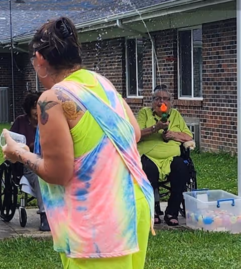 An outdoor scene at a nursing and rehabilitation center showing a woman in a colorful tie-dye tank top and neon green shirt holding a water balloon, facing an elderly person in a wheelchair who is aiming a toy water gun. They are on a grassy area near a brick building with windows. A plastic container filled with water balloons is on the ground nearby.