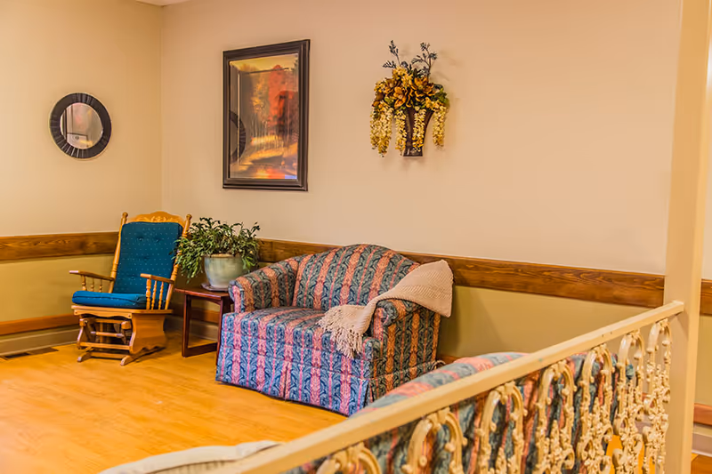 A cozy sitting area in a nursing facility featuring a patterned loveseat with a beige throw blanket draped over one arm, a wooden rocking chair with blue cushions, a small wooden side table with a potted plant, a round wall mirror, a framed landscape painting, and a decorative wall-mounted floral arrangement. The room has light-colored walls with wooden trim and a light wood floor.