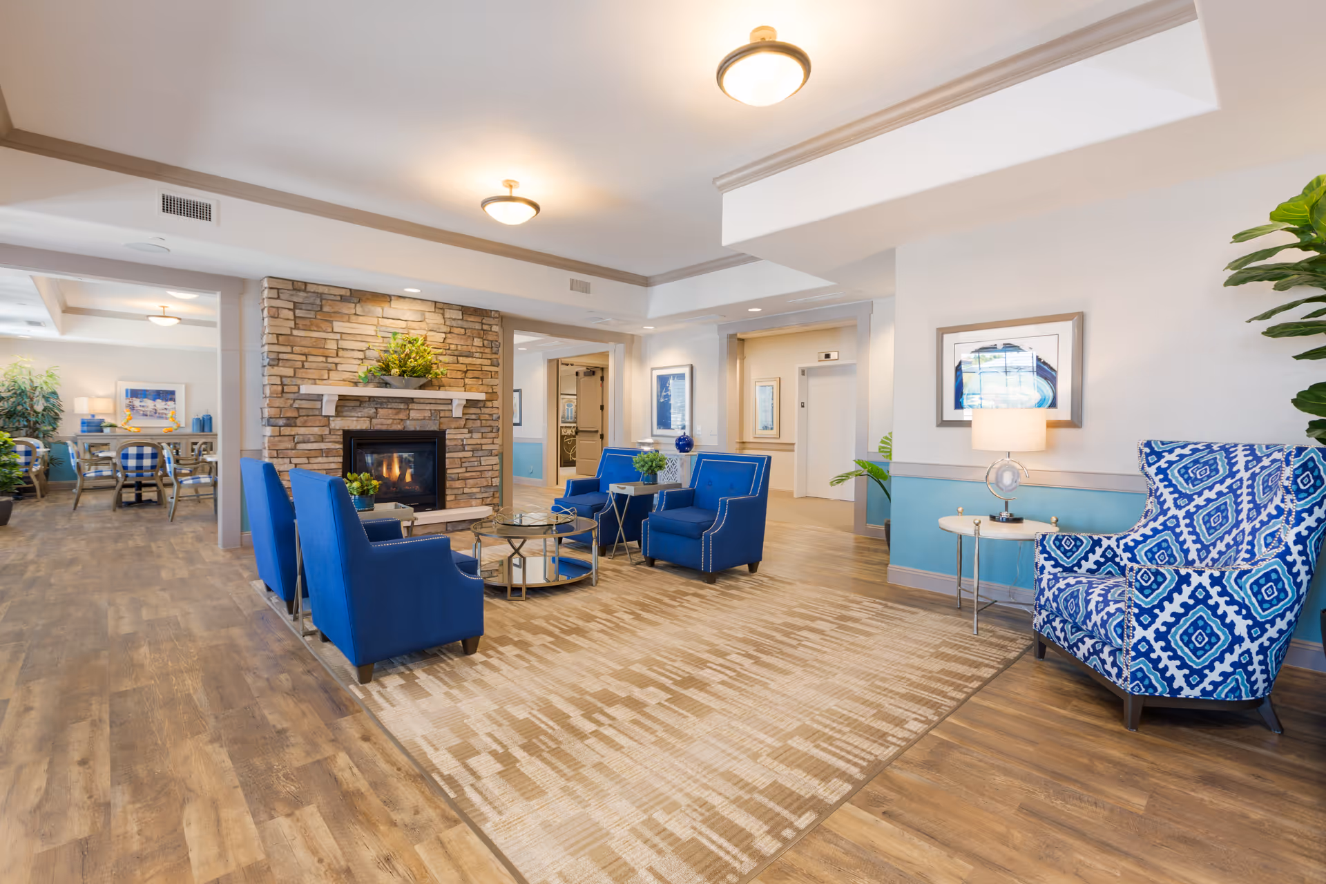 A bright and spacious senior living facility common area featuring a stone fireplace with a mantel decorated with greenery. The seating area includes four blue armchairs arranged around a glass coffee table on a patterned carpet. To the right, there is a blue and white patterned armchair next to a small round table with a lamp. The room has wood flooring, recessed lighting, and light-colored walls with blue wainscoting. In the background, a dining area with tables and chairs is visible.