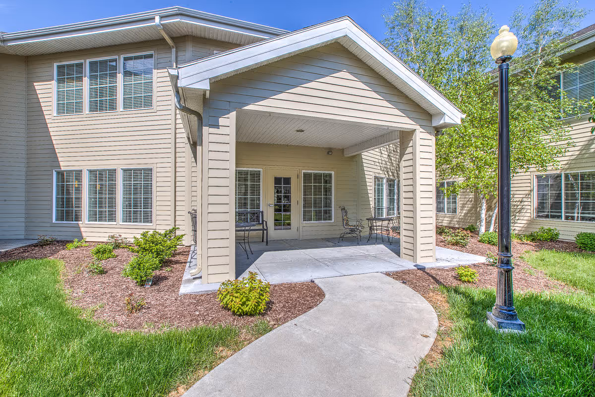 Covered entrance with a small patio and walkway leading to the front of a two-story beige building with landscaping and a lamppost.