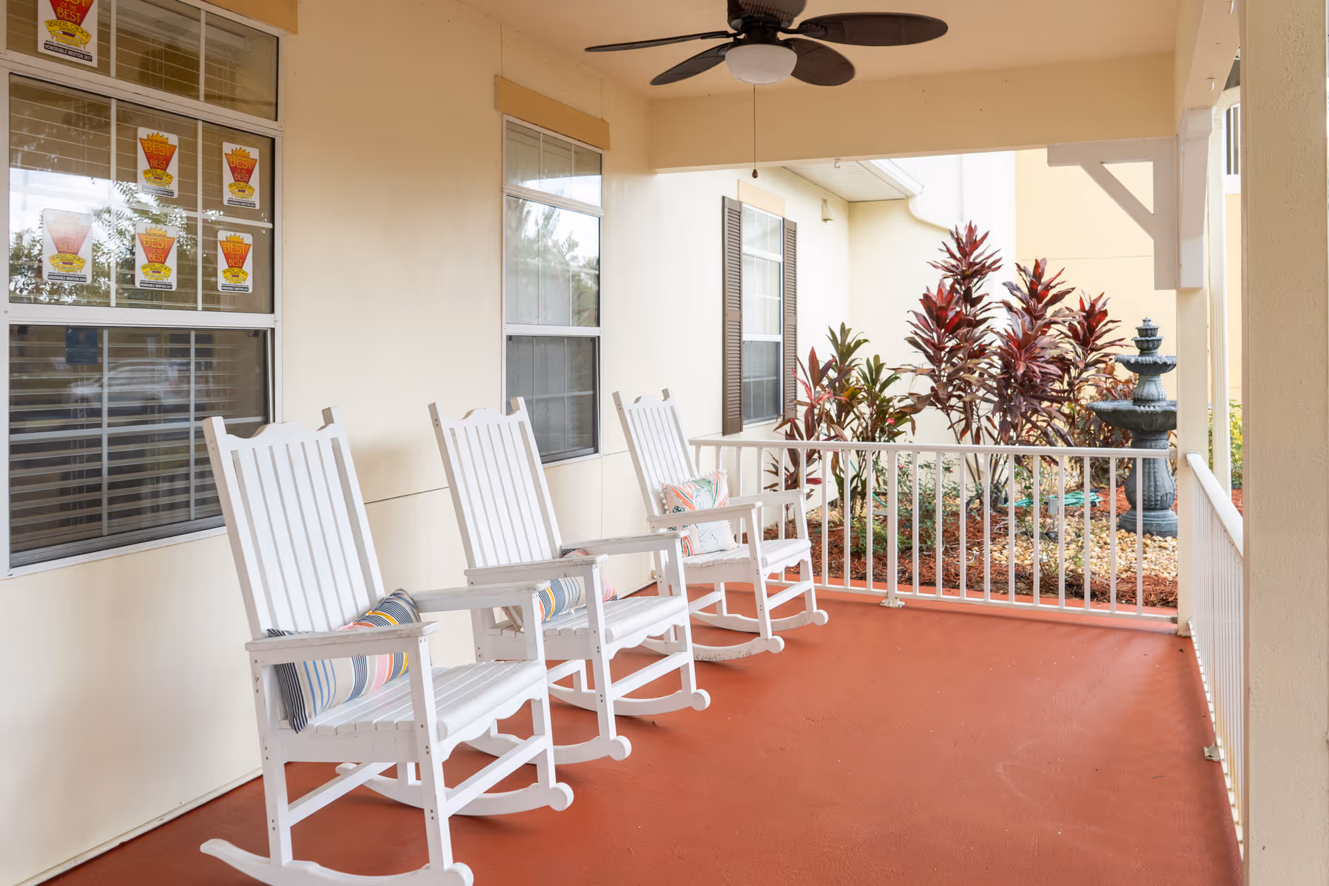 A covered outdoor porch area with four white wooden rocking chairs, each with a colorful cushion. The porch has a red floor, beige walls, two windows with blinds, a ceiling fan, and a white railing. Outside the railing, there are plants and a decorative water fountain.